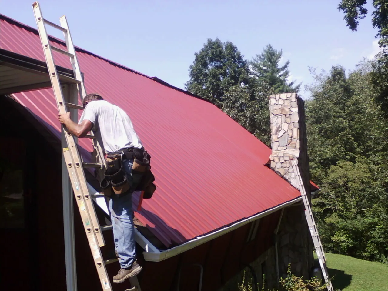 Roofer on a ladder beside a red metal roof and stone chimney — Soffit & Fascia Repair by Roofing Brothers in Venango